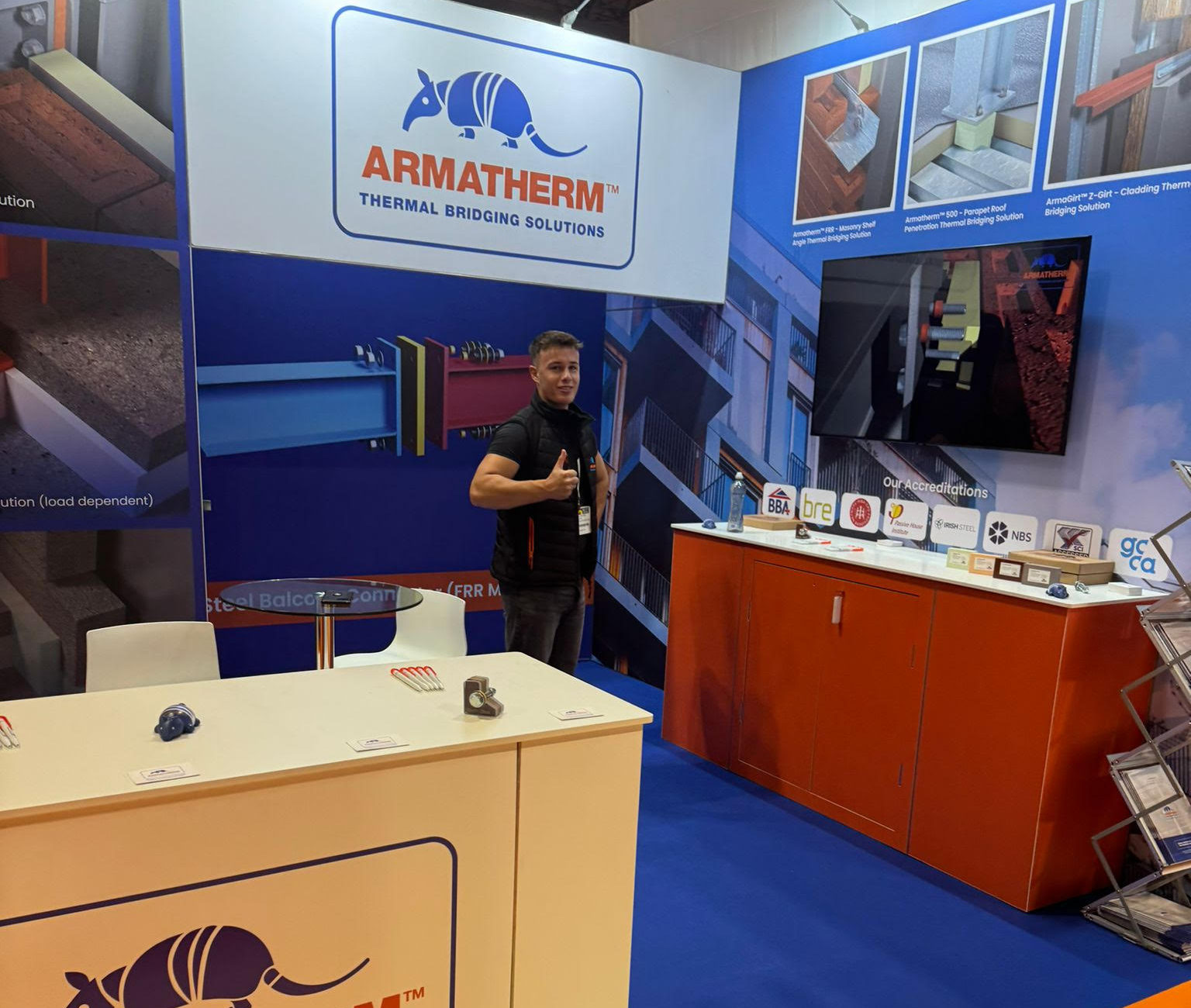 Armatherm staff member giving a thumbs-up at a trade show booth, with product samples, certifications, and a Net Zero Trail floor graphic in view.