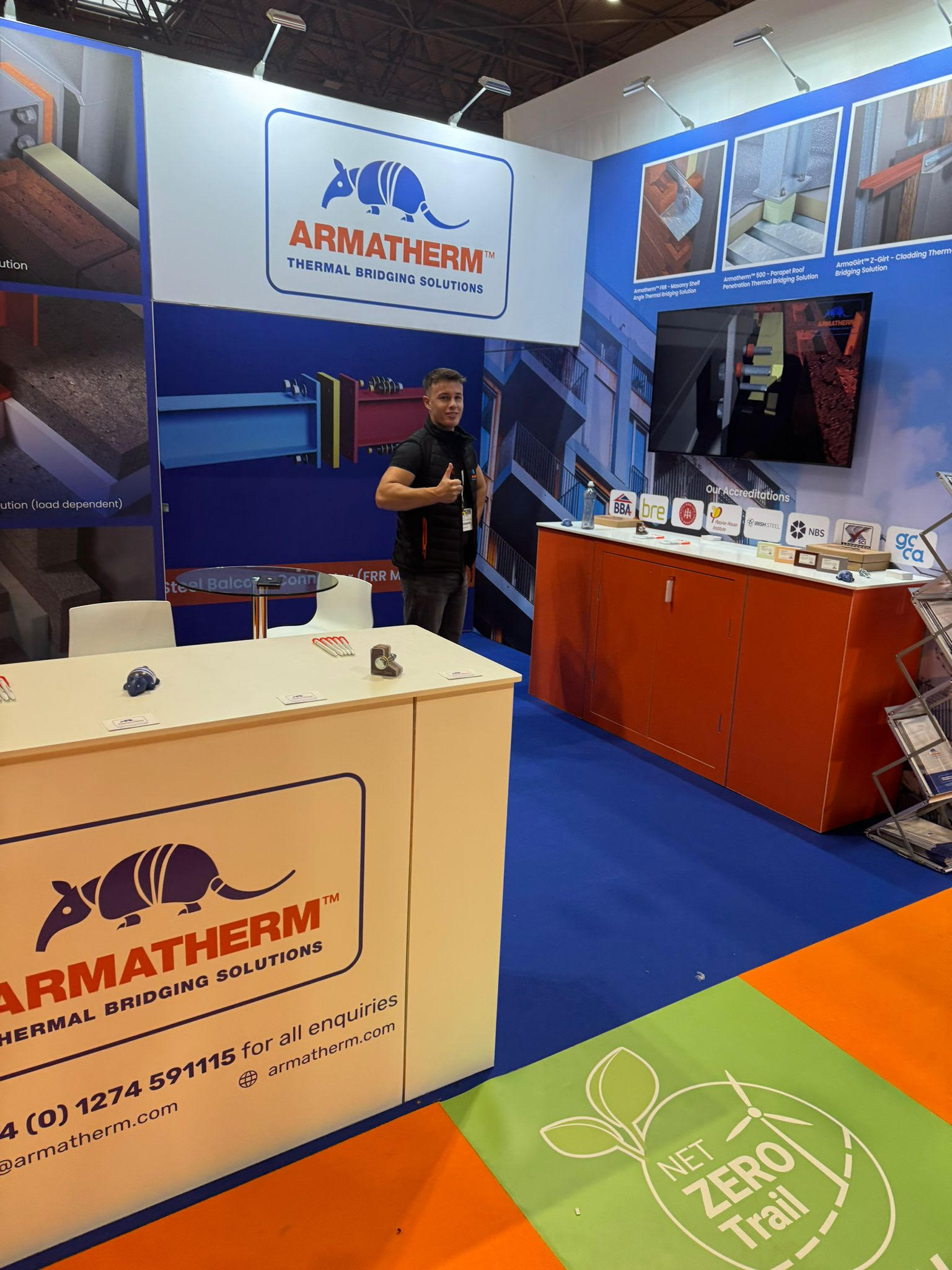 Armatherm staff member giving a thumbs-up at a trade show booth, with product samples, certifications, and a Net Zero Trail floor graphic in view.