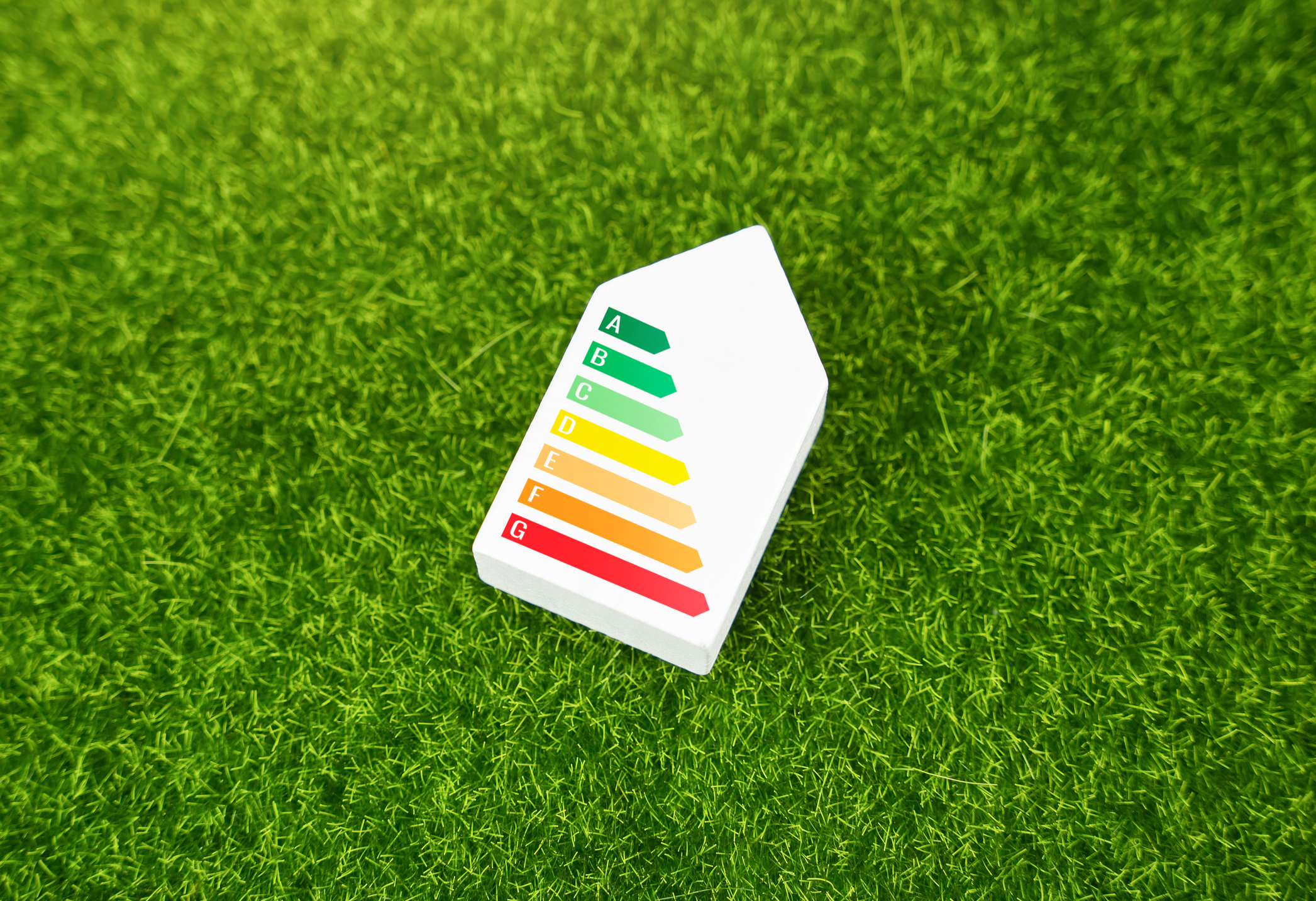 White model shaped like a house displaying an A to G energy rating scale, placed on bright green artificial grass to symbolise energy efficiency and sustainability.