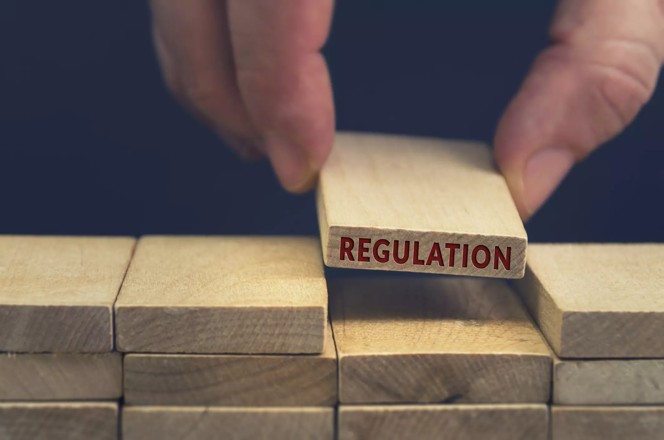 Hand placing a wooden block labeled "Regulation" on top of a structure made from wooden blocks