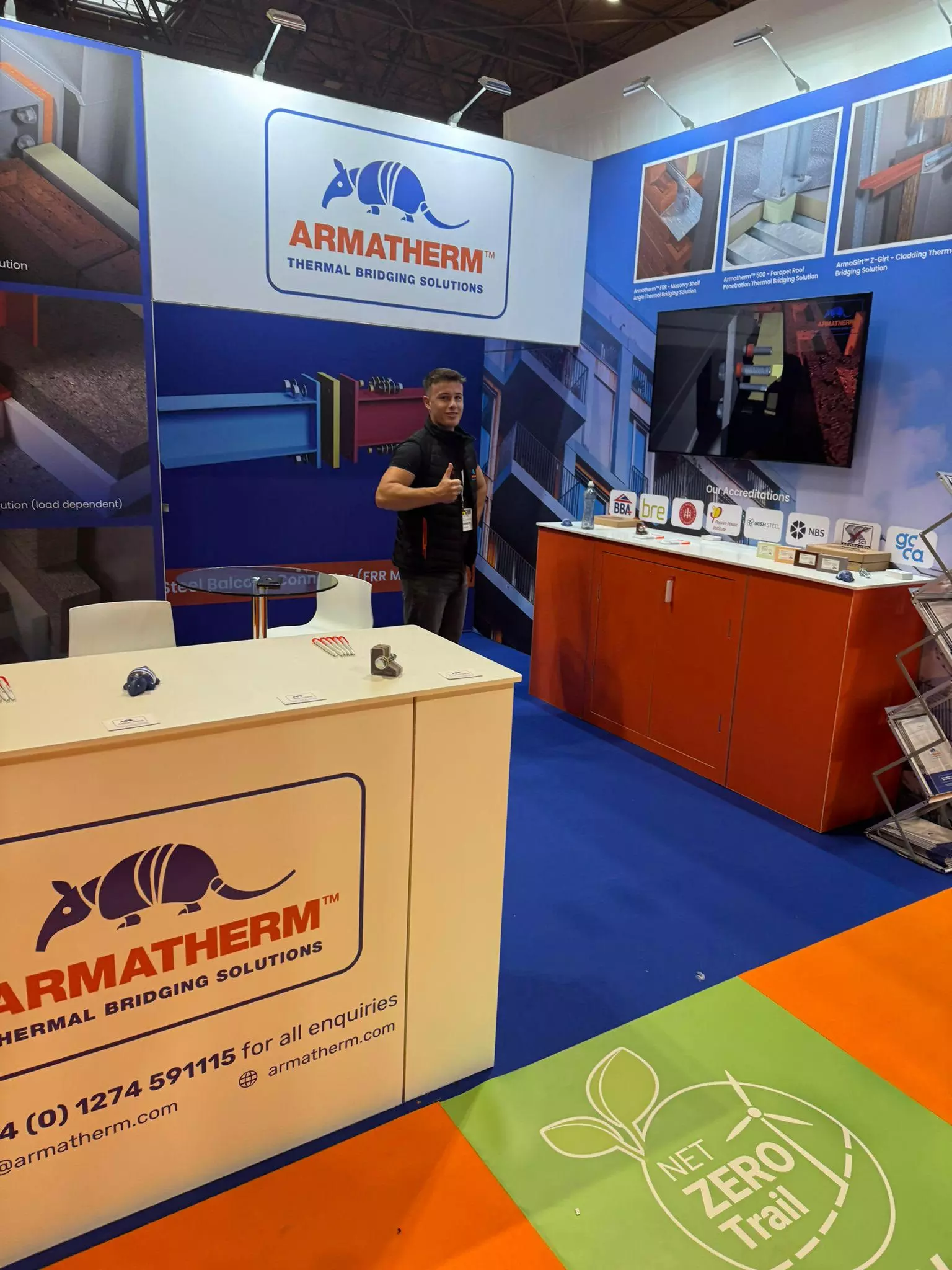 Armatherm staff member giving a thumbs-up at a trade show booth, with product samples, certifications, and a Net Zero Trail floor graphic in view.
