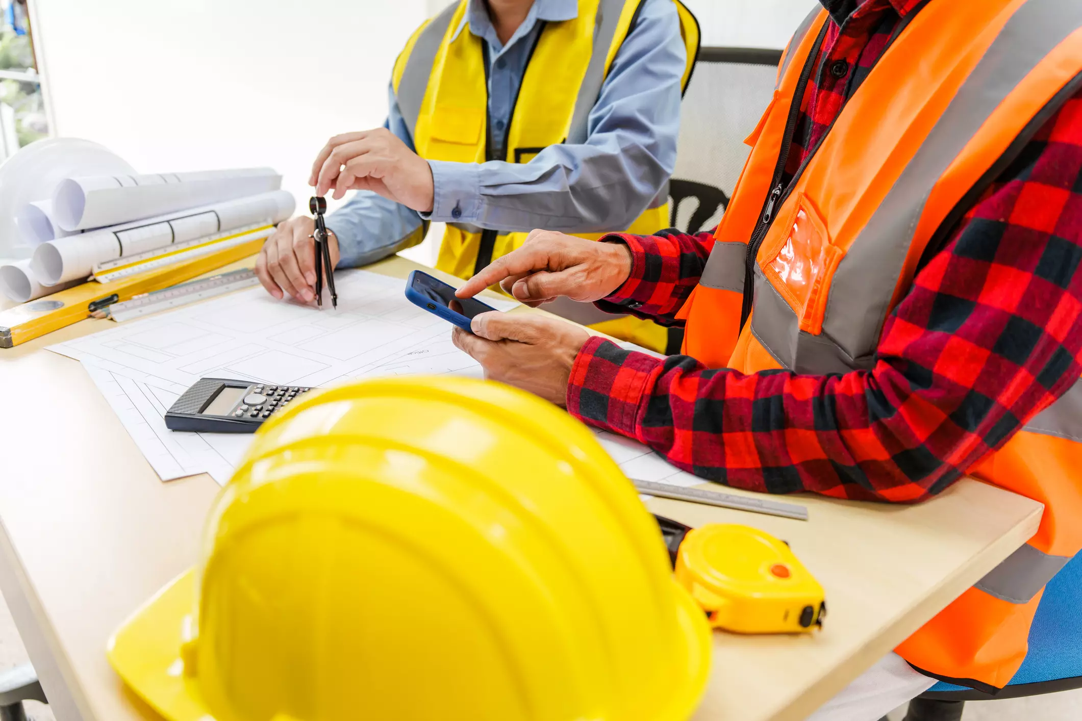 Two construction professionals wearing hi-vis vests working over technical drawings, using a calculator, mobile phone, and compass, with safety helmets and drafting tools on the table.